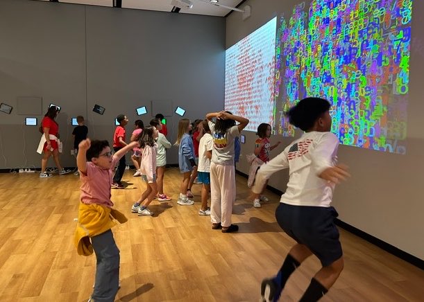 Children energetically dancing and moving around in a room with colorful digital projections on the walls, engaging in an interactive art installation.