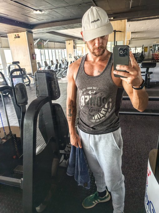 A man taking a mirror selfie in a gym, wearing a gray tank top, gray sweatpants, green sneakers, and a beige cap, holding a phone and a blue towel.