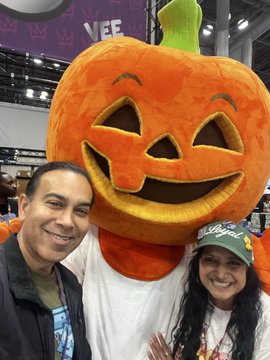 A man and a woman wearing a green cap pose for a selfie with a person in an inflatable pumpkin head costume at what appears to be a convention, with 'VEE' visible in the background.