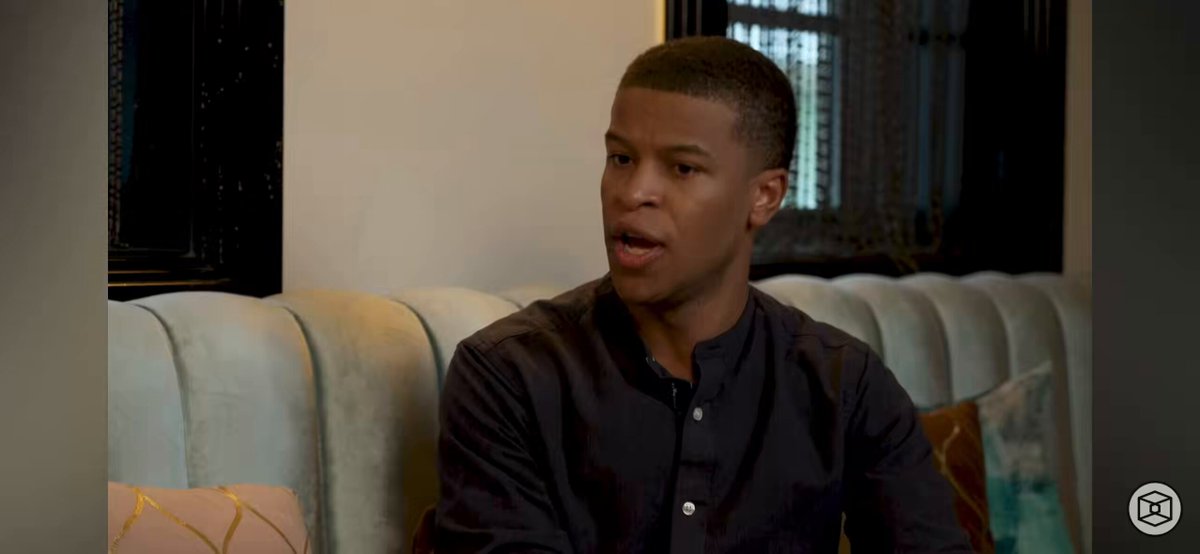 In this image, a man with short hair is sitting down on a couch or chair, wearing a black shirt. He appears to be looking off into the distance while talking on the phone. The background of the photo features a window that allows natural light to enter the room and a bed in the corner. The image also contains text elements such as "Listen closely @XDCNetwork Head of Institutional Travis John" and "Growth pattern year over year will be up and to the right".