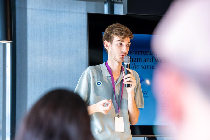 A young man in a gray polo shirt speaks into a microphone at an event, with a screen behind him reading '...currency, chain and Web3 the same.'