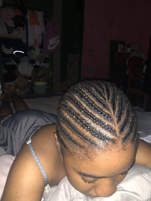 A young woman lies on a bed showing off an intricate braided hairstyle with a leaf-like parting pattern, wearing a grey tank top in a room with a closet and Nike bag visible in the background.