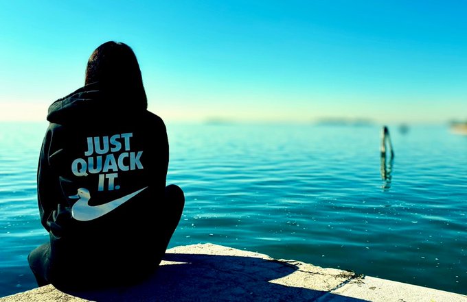 The image features a person sitting alone by the water, wearing a black sweatshirt with white text that says "Just Quackers" written in white letters. The background of the photo shows a beautiful view of the ocean or sea, which adds to the overall atmosphere and setting of the scene.