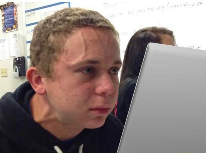 The image shows a young man sitting at a desk, looking down at his laptop computer screen with a sad expression. The background of the photo includes a whiteboard or bulletin board, which could be part of an educational setting like a classroom. There is also another person in the background who appears to be looking at something on the same whiteboard as the young man.