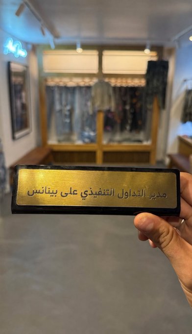 A hand holds a gold nameplate reading "مدير التداول التنفيذي علي بينانس" (Executive Trading Director Ali Binance) inside a clothing store, humorously connecting the user's playful jab about an "upgrade from yourself on Binance" to the Arabic nameplate featuring their own name paired with the exchange.