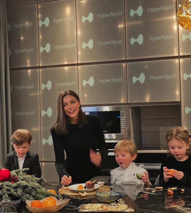 A woman and three children in a kitchen decorated with 'Hyperliquid' logos on the cabinetry, preparing food with various ingredients spread out on the counter.