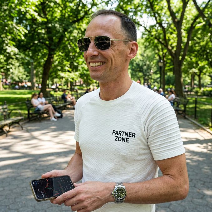 The image shows a man wearing sunglasses, holding his cell phone with both hands, smiling at the camera. The background of the photo features a park setting with several people sitting on benches or standing around. There are also trees visible in the scene. The caption for this image reads "GM Your portfolio can grow on Saturday while you enjoy a walk in the park. Have a great Saturday!"
