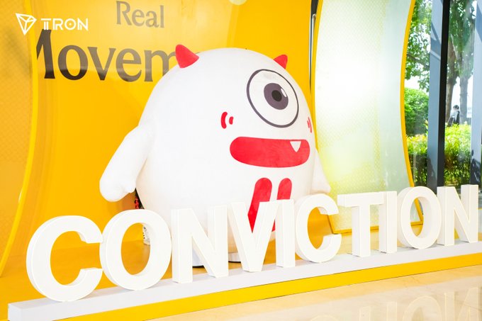 A white and red mascot character stands behind large 3D letters spelling 'CONVICTION', with a TRON logo and partial text 'Real Movem' visible on a yellow background.
