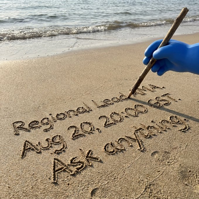 A blue-gloved hand uses a stick to write "Regional Lead AMA, Aug 20, 20:00 GST, Ask Anything" in the sand at the edge of the sea, visually echoing the casual, open-ended invitation for the upcoming live Q&A session.