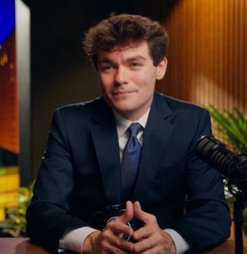A young man in a dark navy suit and blue tie sits at a desk with a microphone, appearing to be a TV show guest or host.