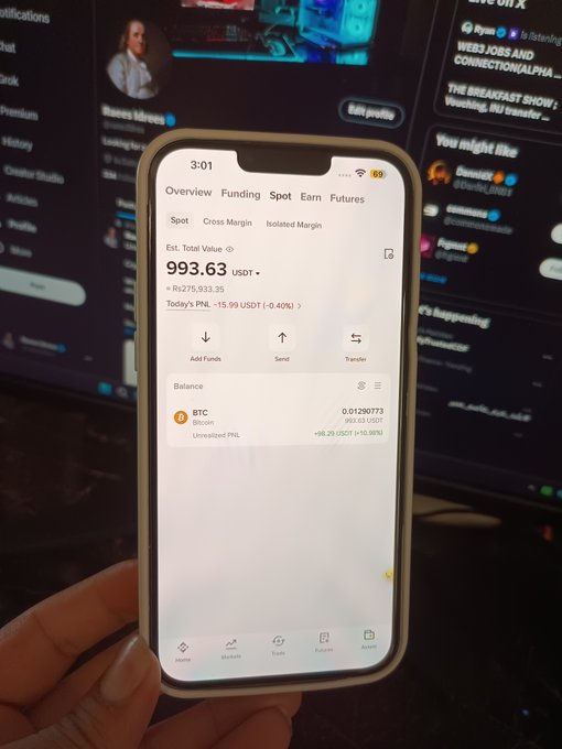 A hand holds a smartphone displaying a crypto trading app showing '993.63 USDT' total value with a BTC balance, set against a blurred background of a trading interface.