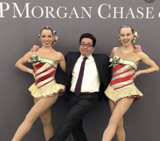 A man in a suit and glasses poses with two showgirls in candy-cane striped costumes in front of a JPMorgan Chase backdrop.