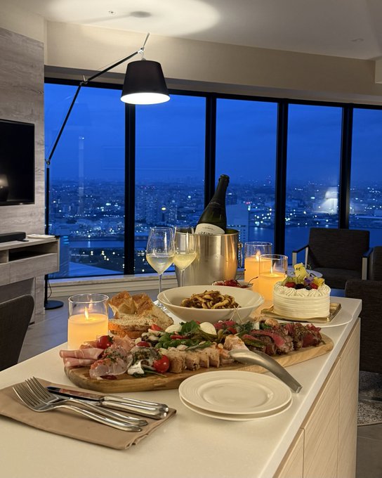 An elegant dinner spread featuring a charcuterie board with meats, cheeses, and vegetables, accompanied by wine, champagne, and candles, set against a stunning cityscape view through floor-to-ceiling windows at twilight.