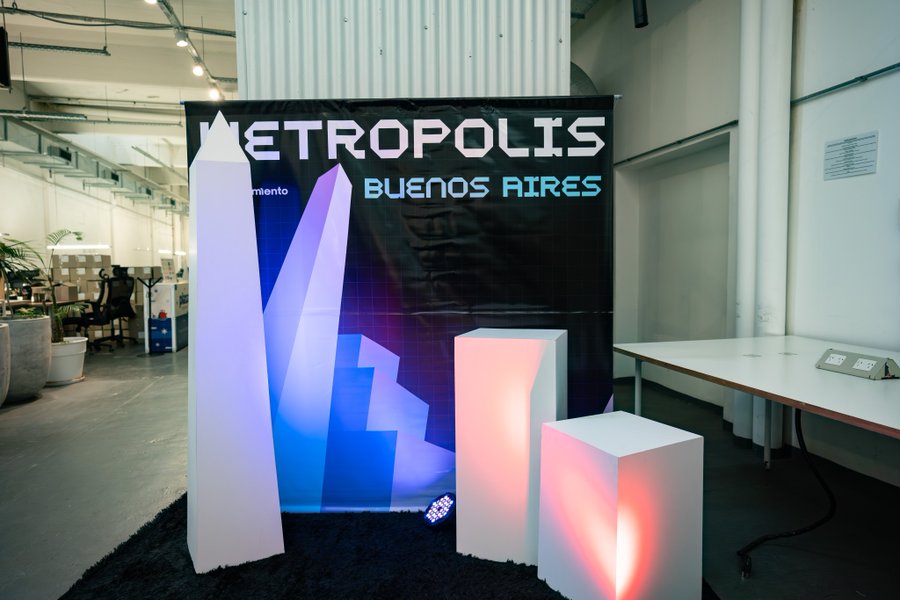 An indoor event space featuring a 'METROPOLIS BUENOS AIRES' banner on a dark backdrop, accompanied by illuminated white geometric pillars and cubes glowing in blue, pink, and red hues.