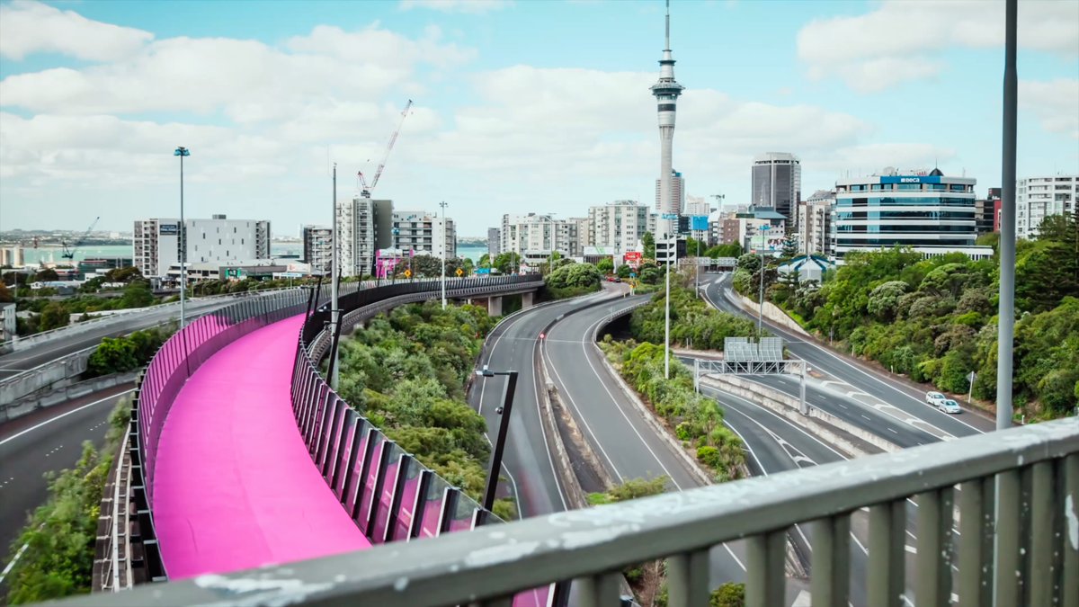 A cityscape of Auckland, New Zealand, featuring the Sky Tower, highways, urban buildings, and a distinctive pink curved structure in the foreground.