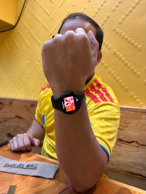 The image features a man sitting at a table with his arm raised, wearing a watch that has an orange face. The background of the photo shows a yellow wall, which complements the man's attire. The caption of the image reads "Hoy juega la Sele en Mexico! Y con @MetaMask Card puedes viajar y pagar en cualquier lugar del mundo usando tus dólares digitales". This post seems to be promoting a travel-related hashtag, encouraging people to explore the world using their smartphones and social media platforms.

The man's watch has an orange face, which contrasts with his yellow shirt. The image also includes a wooden table in the background, adding a rustic touch to the scene.