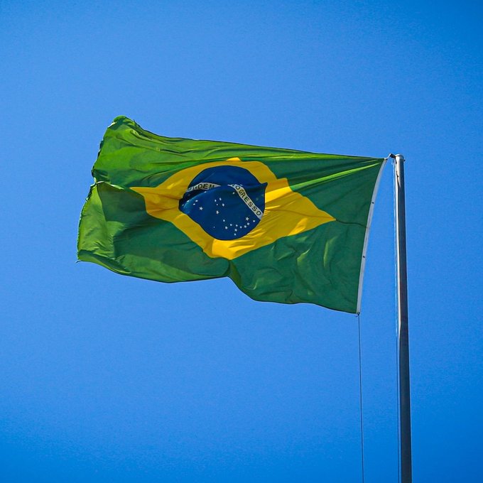 The Brazilian flag waves prominently against a clear blue sky, featuring its iconic green field, yellow rhombus, and blue celestial globe inscribed with "ORDEM E PROGRESSO." The image visually anchors news about Brazil's tightening crypto regulations, symbolizing the nation whose capital rules may drastically reduce licensed crypto providers.