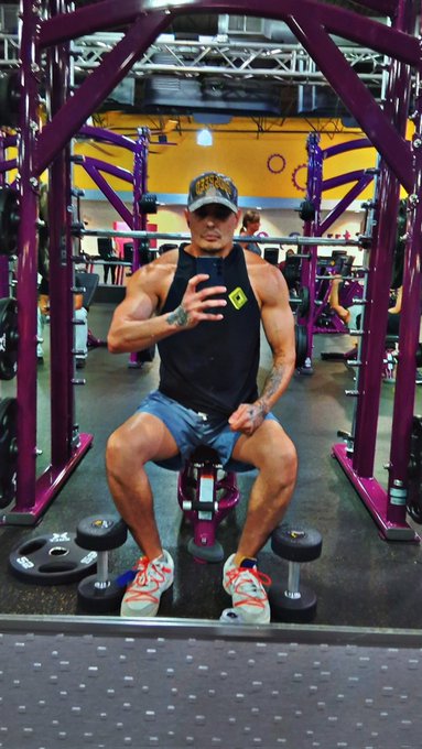 A muscular man wearing a black tank top and cap taking a mirror selfie in a gym with purple equipment.