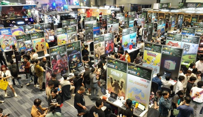 An aerial view of a busy gaming convention/exhibition hall with numerous booths displaying indie video games like Stray, World of Goo, and Strider, with crowds of attendees browsing.