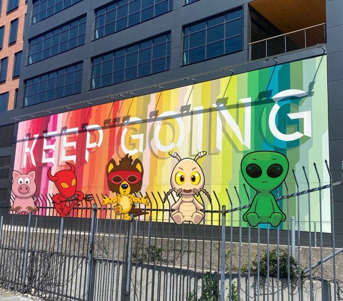 A colorful rainbow billboard on a building displays 'KEEP GOING' along with cartoon characters including a pig, red devil, lion, larva, and alien.