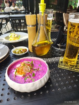 An outdoor restaurant table featuring a bowl of pink soup (likely borscht) garnished with vegetables, a large glass of beer, and an olive oil dispenser.
