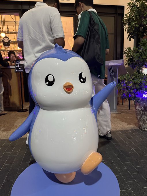 A glossy statue of Piplup, the blue penguin Pokémon, sitting on a blue base with one foot raised, displayed in what appears to be a Pokémon Center shop.