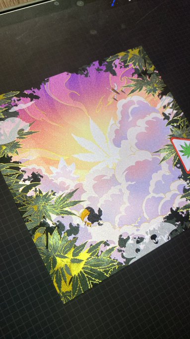 A colorful artwork on a cutting mat featuring a pastel sky in pink and orange, white clouds, green palm/cannabis leaves, and a large white cannabis leaf, with a small bee figure.