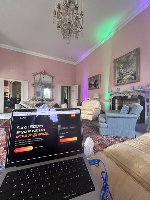 A MacBook displaying a 'Send USDC to anyone with an email or @handle' webpage sits on a person's lap in an ornate room with pink walls, a crystal chandelier, and colorful LED lighting.
