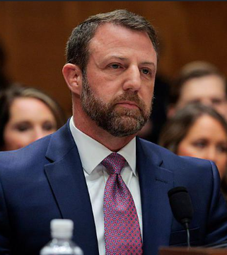 The image shows a man wearing a suit, tie, and beard, standing at a podium with his hands behind his back. He appears to be giving a speech or presentation. The caption reads "What the hell did DOGE even do???". This suggests that there might have been an incident involving a dog in the room where the event took place, possibly causing some disruption or distraction during the proceedings.

The man's facial expression and body language indicate that he is serious about addressing the issue at hand. The presence of a water bottle on the podium further supports this notion, as it could be used to quench his thirst while he speaks.