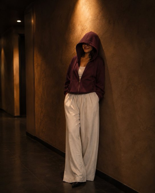 A woman in a maroon zip-up hoodie and white wide-leg pants leans against a textured wall in a dimly lit hallway.