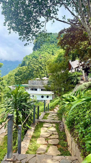 A stone pathway with a rope railing leading down toward a white building with round windows, nestled among lush green mountains.