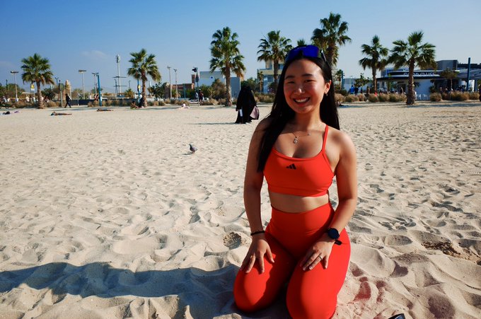 Paris kneels on a sandy beach in a bright red Adidas workout set, smiling warmly with sunglasses pushed up on her head against a backdrop of palm trees and a sunny, boardwalk-lined shore.

This casual, active look fits the post's reintroduction, capturing the vibrant, wellness-focused energy of someone who curates experiences and brings people together.