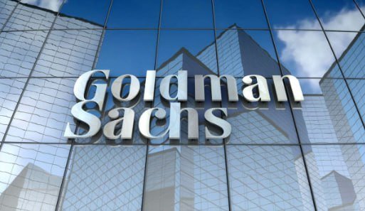 The 'Goldman Sachs' logo displayed on the reflective glass facade of a modern building, reflecting the blue sky and clouds.