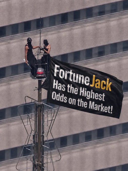 Two people stand on top of a traffic light pole unfurling a large black banner reading "FortuneJack Has the Highest Odds on the Market!" in white and yellow text, overlooking a modern high-rise building.

The image's bold advertising banner for FortuneJack's betting odds serves as a striking visual hook, tying into the post's promotional giveaway of $30 in SOL, where the high-stakes, "highest odds" theme echoes the risk-and-reward vibe of crypto giveaway contests.
