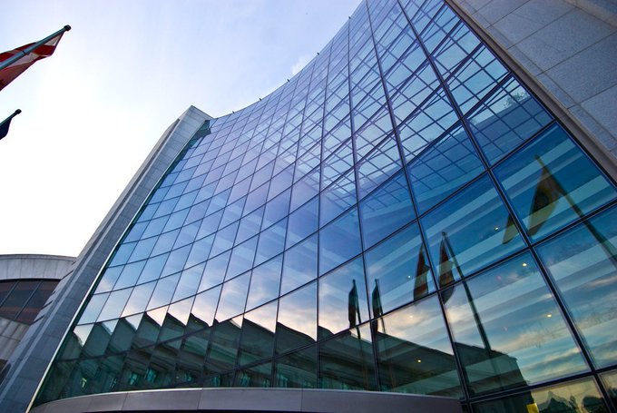 A low-angle view of a large modern building with a curved glass facade reflecting the sky and flags (including what appears to be a Canadian flag), with another flag visible on the left.
