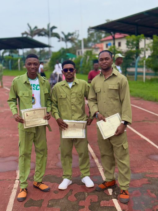 Three young men in olive-green NYSC-style uniforms proudly hold up their certificates on an outdoor running track, with one displaying a "1" on his document, celebrating the completion of their service year as referenced in the caption's mention of a closed chapter opening another—likely the end of their National Youth Service Corps program.