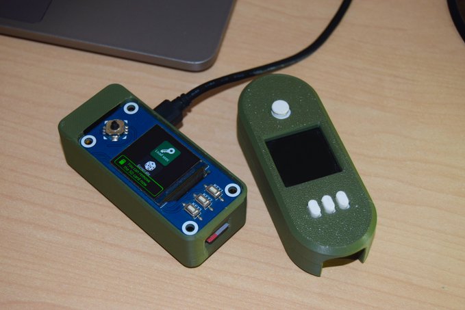 Two green electronic devices with USB cables on a wooden desk, one open showing a small OLED screen with a loading icon and circuit board, the other closed with a blank screen and three small buttons.
