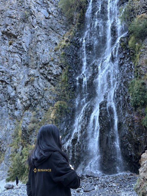 A person with long black hair wearing a black jacket with the yellow 'BINANCE' logo stands with their back to the camera, viewing a large waterfall cascading down a rocky cliff.