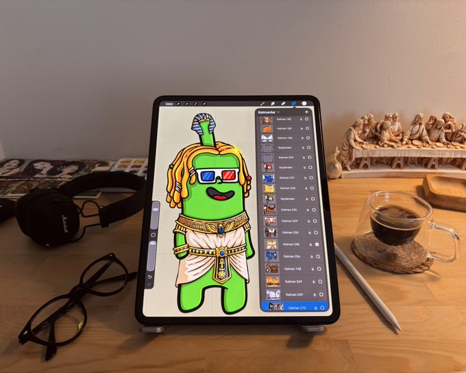 An iPad displayed on a wooden desk shows a digital illustration in progress featuring a green cartoon character dressed as an Egyptian pharaoh with 3D glasses, alongside a layer panel showing various "Kalman" assets. The scene, complete with a coffee cup, headphones, and reading glasses, visually supports the creator's statement that the artwork is entirely hand-drawn from scratch, showcasing the original digital creative process behind the collection.