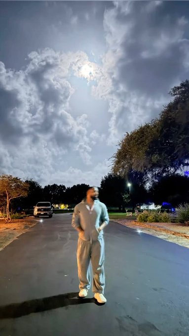 A person in a gray tracksuit stands in the middle of a moonlit street, casting a long shadow under a dramatic, cloudy night sky, embodying the casual "gm" (good morning) greeting shared with fellow crypto and social media users. The serene nighttime setting paired with the relaxed pose captures the laid-back spirit of the "gm" community ritual.