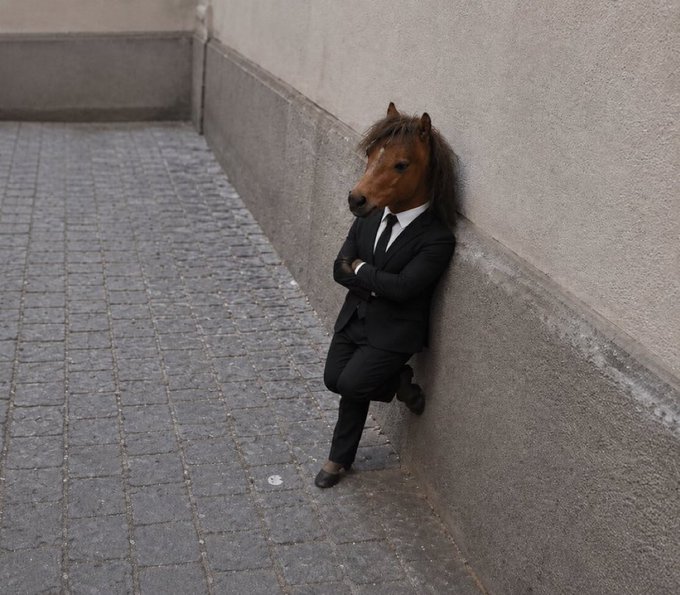 A horse head in a black business suit leans nonchalantly against a stone wall on a cobblestone street, embodying the unbothered "$HORSE" meme mascot known for its deadpan indifference. The image visually captures the "Nonchalant Horse" persona referenced in the post, depicting the stoic, suit-wearing equine figure that's become a viral symbol of market calm amid chaos.