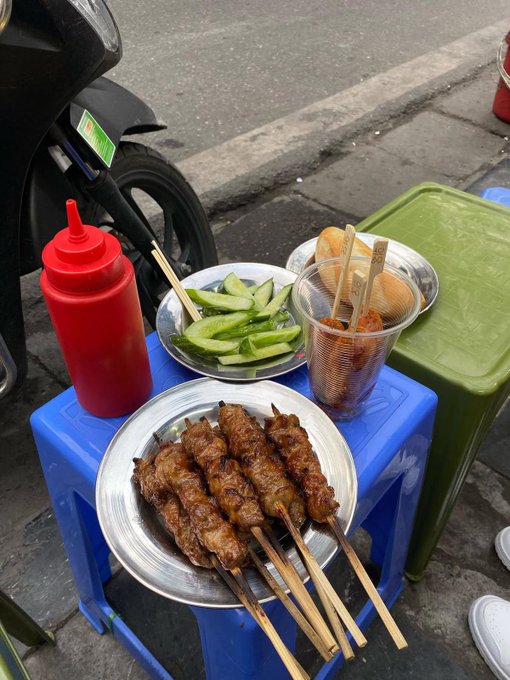 Vietnamese street food setup on blue plastic stools, featuring a plate of grilled meat skewers (thịt nướng), a bowl of sliced cucumbers, a cup of dipping sauce, bread, and a red squeeze bottle of chili sauce, with a motorbike in the background.