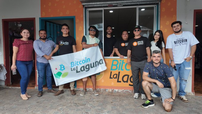 A group of smiling people posing for a photo holding a "Bitcoin La Laguna" banner, standing in front of a brightly colored orange and yellow storefront.