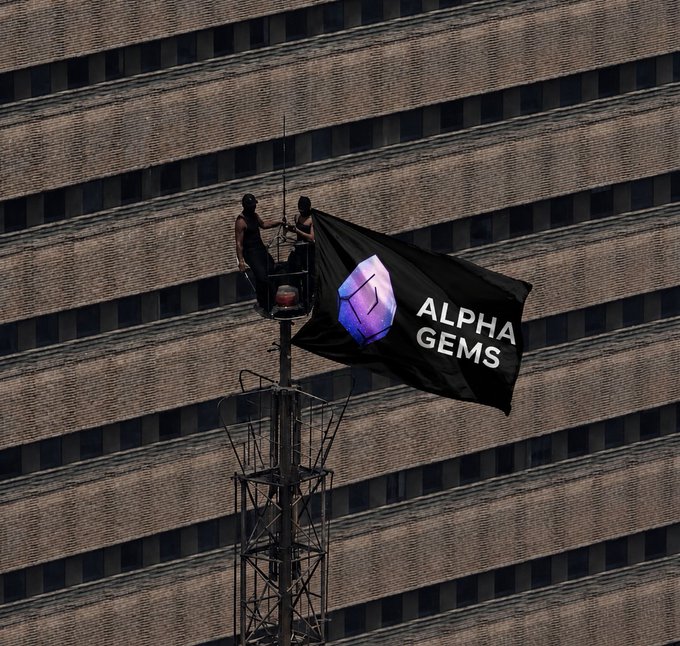 Two people in a lift attach a black flag reading 'ALPHA GEMS' with a purple cube logo to a tall structure against a brick building.