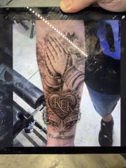 A tattoo stencil of praying hands and a sacred heart with "MOM" is visible on the customer's arm, with the artist's hand hovering above, ready to begin inking. The "Stencil is on" caption refers to this preparatory step before the actual tattooing begins.