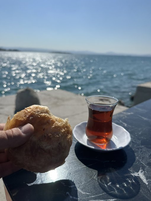 A hand holds up a piece of fried dough or bread alongside a classic tulip-shaped glass of dark Turkish tea on a saucer, all set on a marble surface with a sparkling blue sea in the background—capturing a serene seaside breakfast moment.
