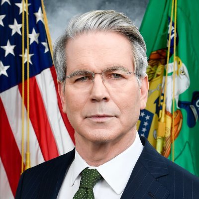 Official portrait of a gray-haired man in glasses, a navy suit, and green tie, standing before an American flag and another flag.