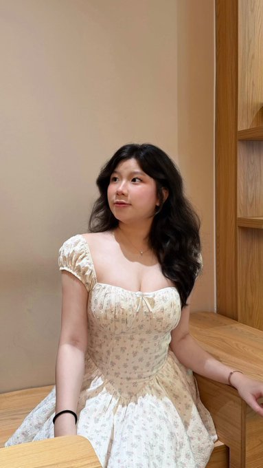 A young woman with long dark hair poses in a cream-colored floral dress, looking off to the side while seated near a wooden shelf.