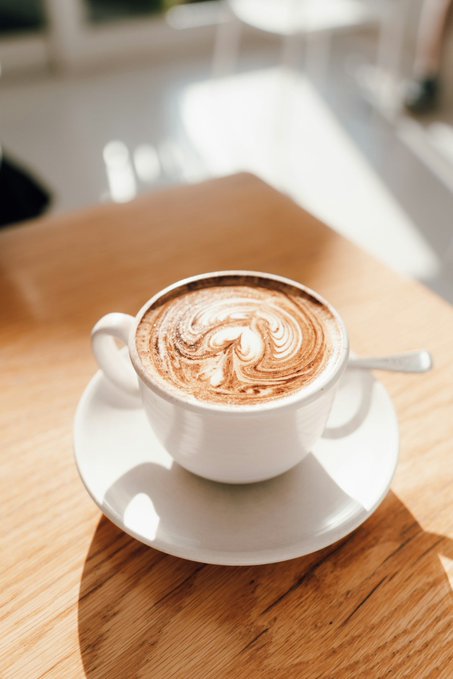 A cappuccino with intricate latte art sits on a wooden café table in warm sunlight, paired with a contemplative post suggesting the poster believes Bitcoin ($BTC) has likely reached a local top. The image's serene coffee moment visually echoes the reflective, wait-and-see sentiment behind the bearish $BTC outlook.