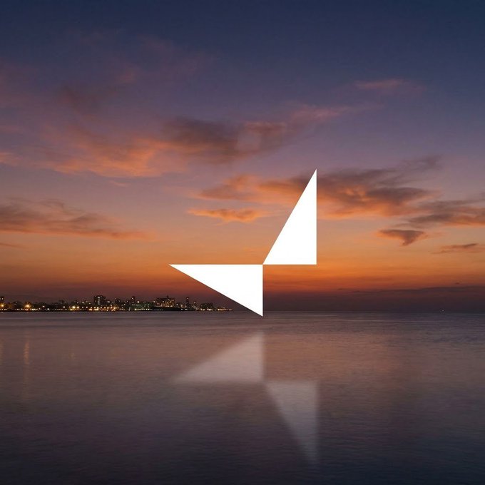 A stylized white geometric logo resembling an angular 'A' is centered over a serene sunset seascape with a city skyline in the distance.